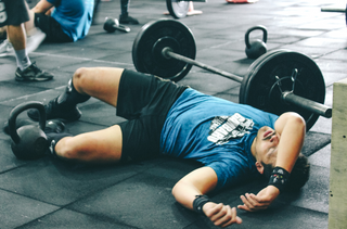 A person lying on the gym floor exhausted after an intense workout, with a barbell and kettlebells nearby, depicting the physical toll of strength training.